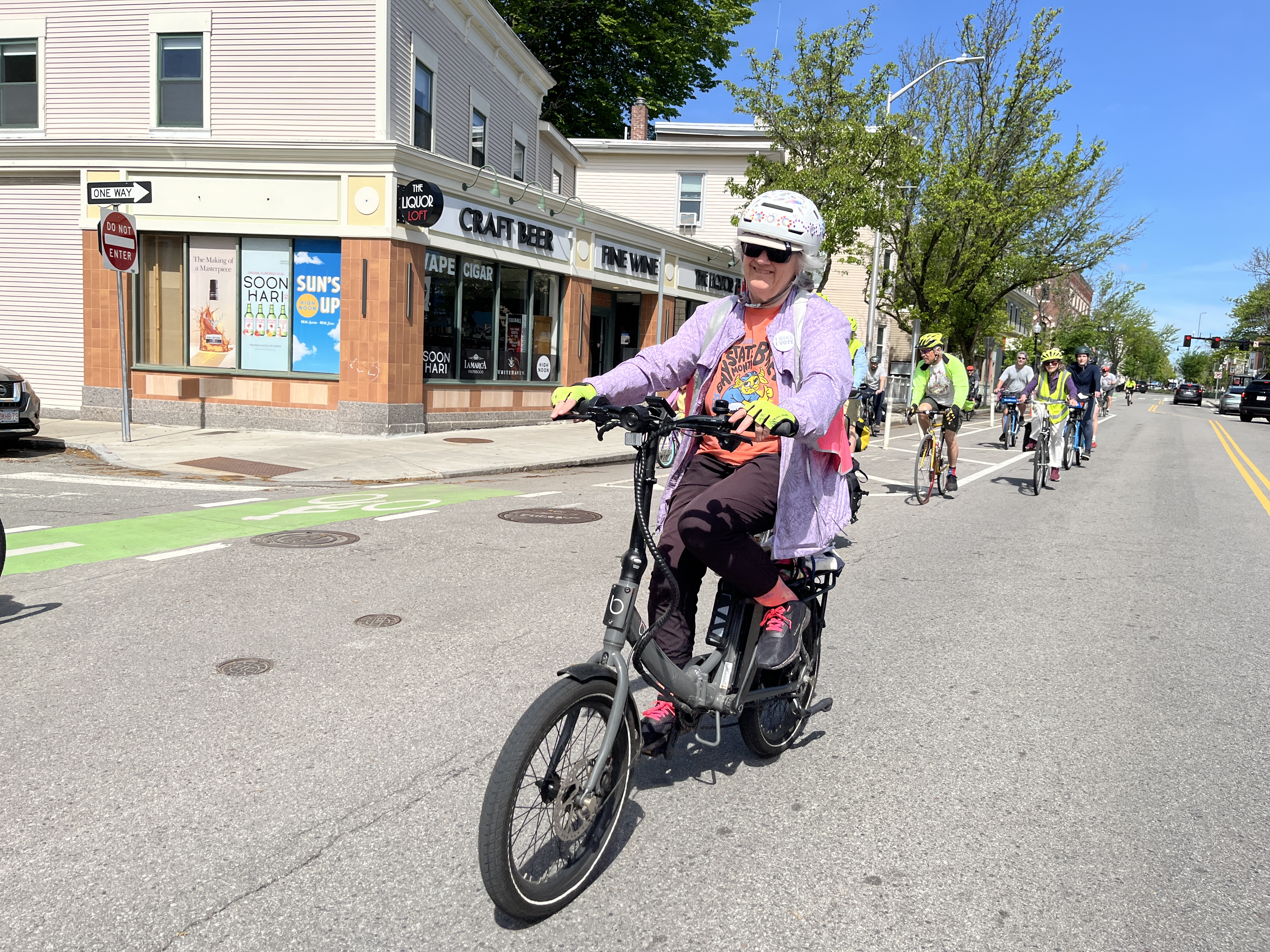A bicyclist rides down the middle of the street past a convenience store. Other bicyclists are visible in the background.