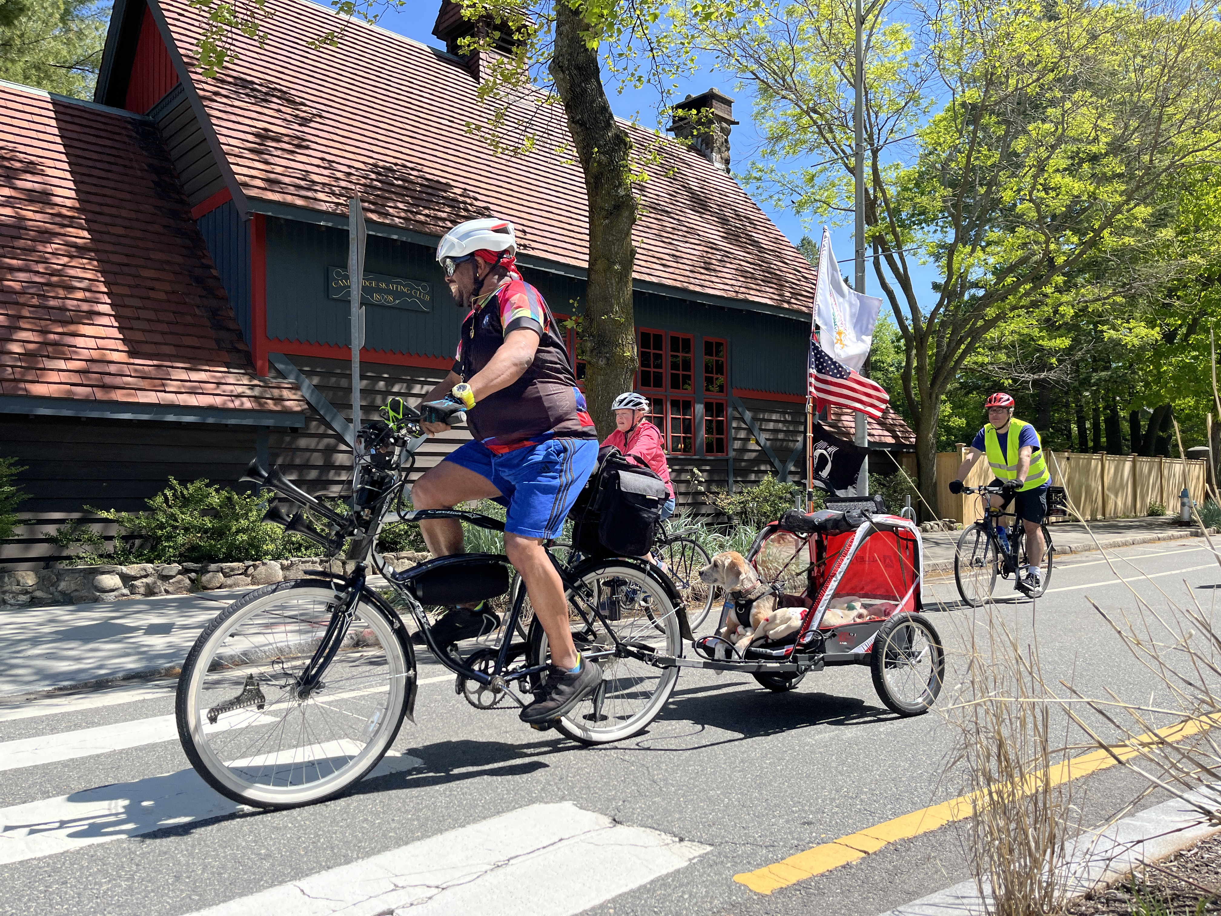 A biker with a dog trailer and flags rides past the Cambridge Skating Club building.