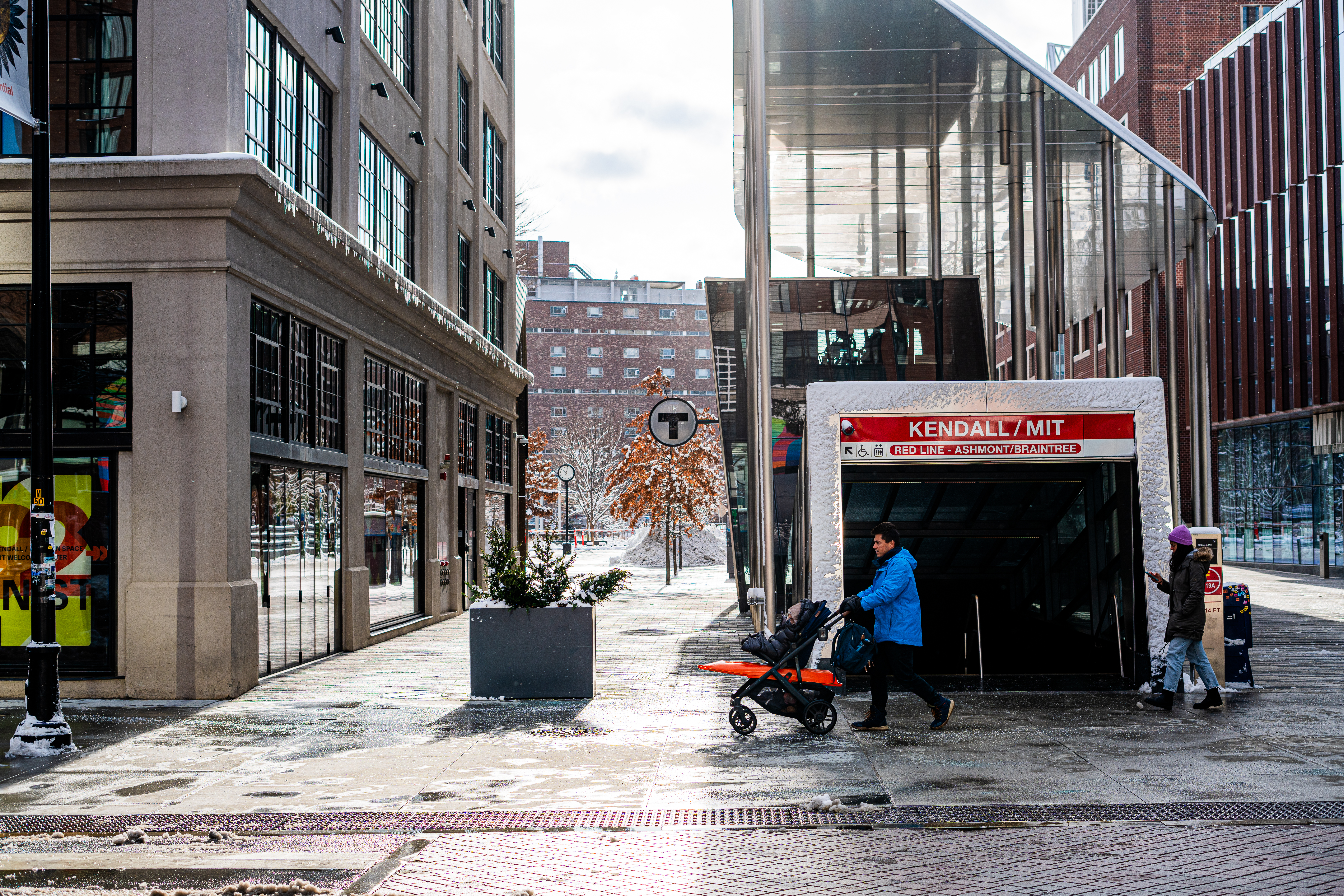 A man pushes a baby carriage past the Kendall Square / MIT T stop.