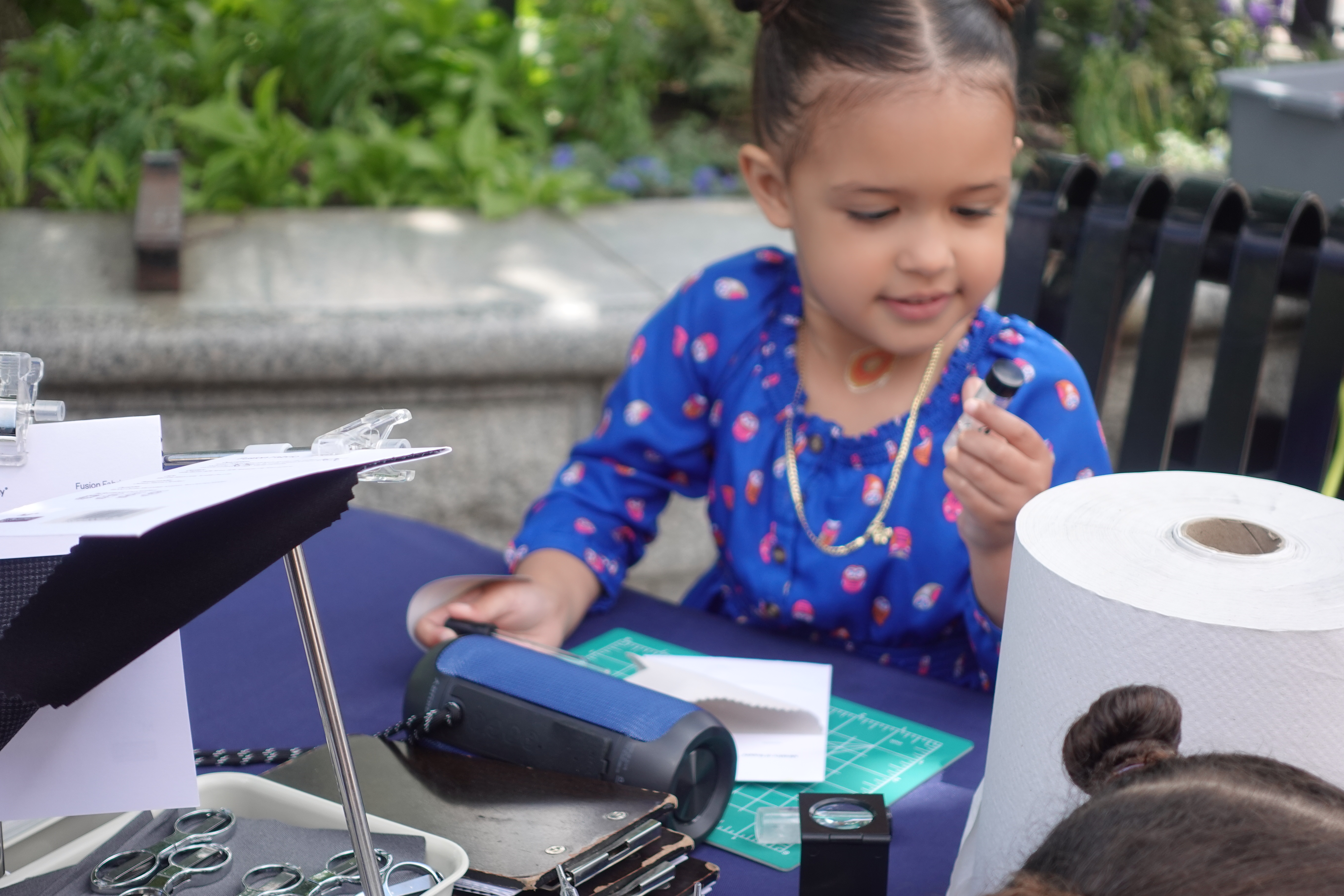child wearing a blue top, seated outdoors at a Cambridge Plays event engaging in a craft activity