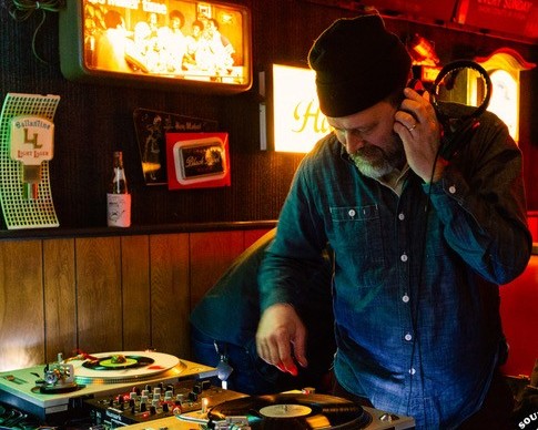 DJ standing at vinyl turntable holding headphones up to their ear. Setting is a dim bar with glowing square lights in the background.