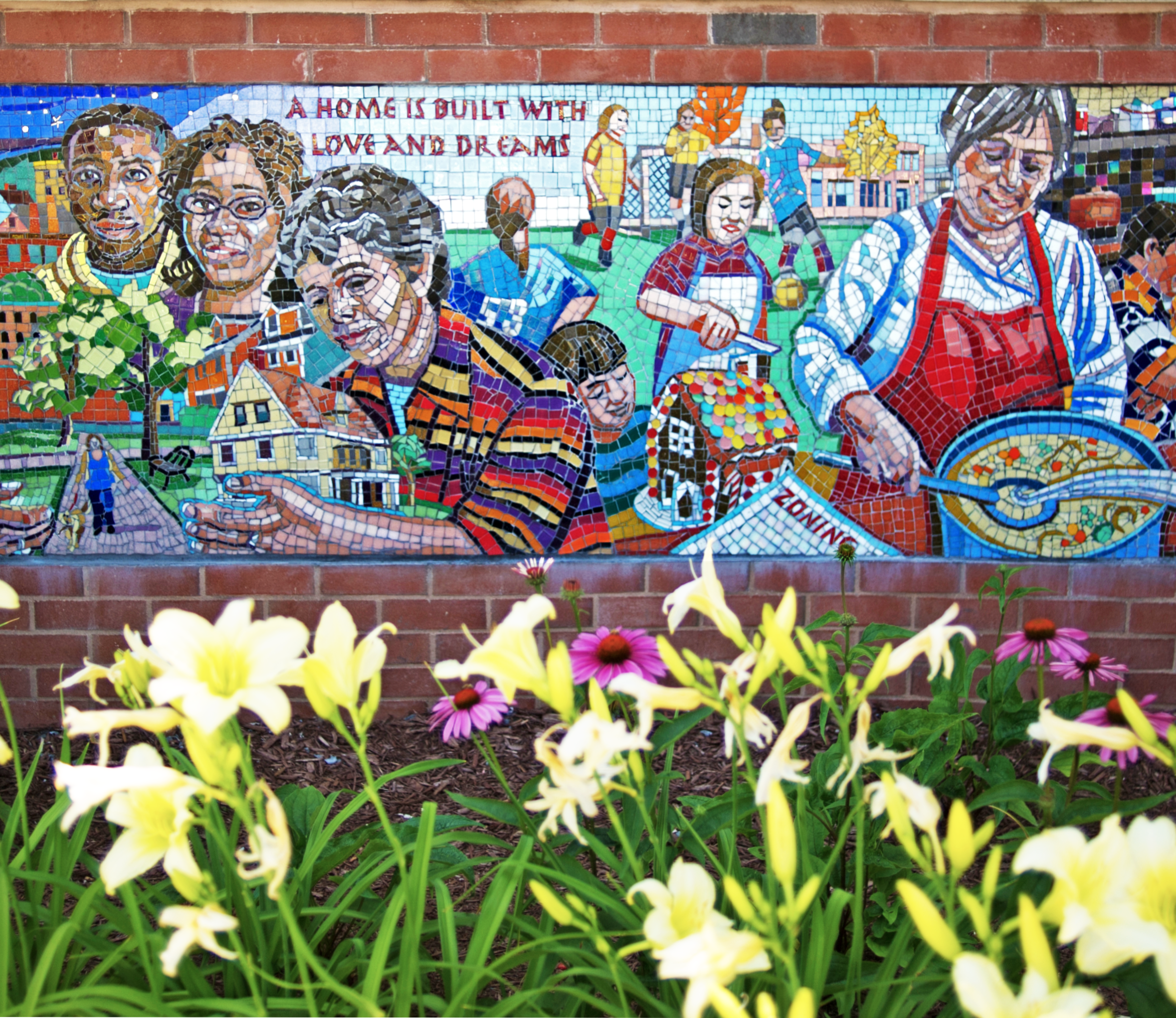 Bed of blooming flowers underneath a colorful mosaic mural of diverse faces, houses, and people gathering and cooking