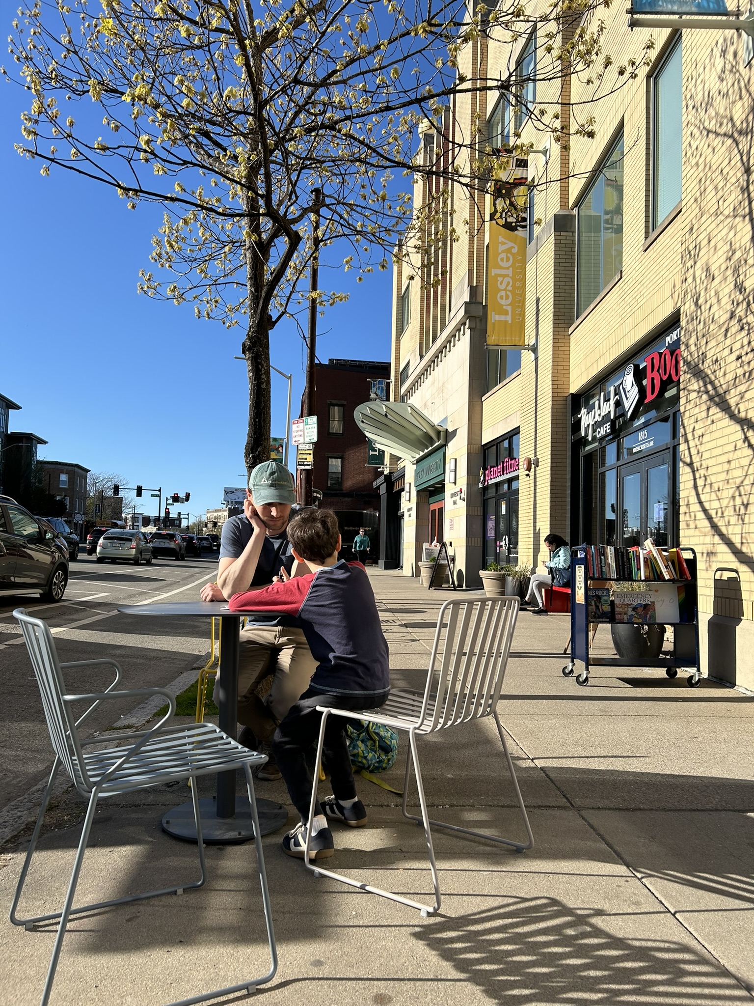 Two people sitting at Cool Spots table and chairs on Mass Ave outside of Lesley University Hall. In the background is a tree with budding leaves and blue sky.