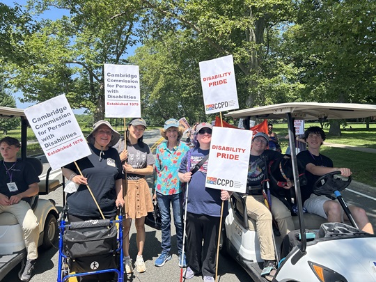 A group of people holding signs are gathered in a Cambridge park to celebrate Disability Pride and march in the 2025 Cambridge Arts River Festival