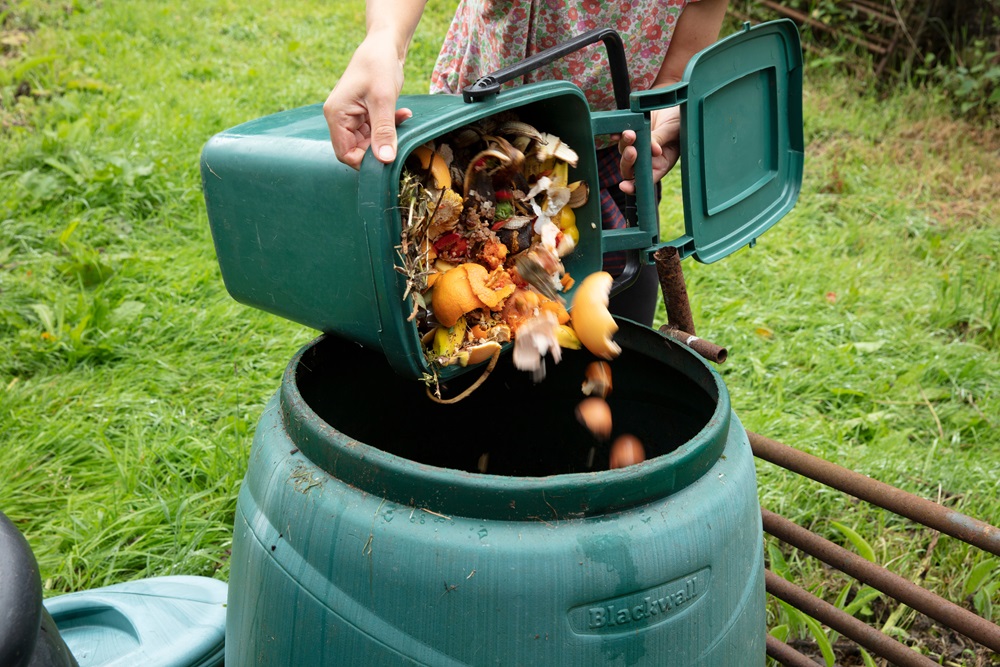A person is emptying a green kitchen compost bin full of food scraps into a larger green compost container outdoors.