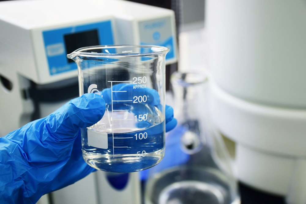 A gloved hand holding a clear beaker filled with water in a laboratory setting with scientific equipment in the background.