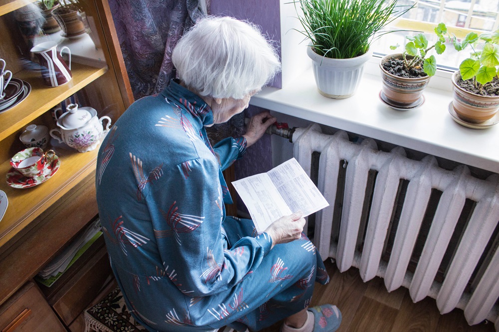 A person is sitting beside a radiator, reading a heating bill by a windowsill with various potted plants and decorative items.