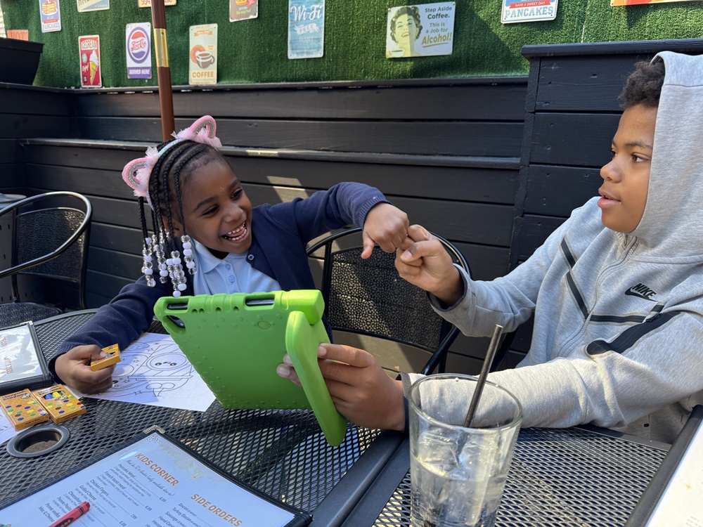 Two children smiling and fist-bumping at a restaurant table. There are art supplies, a menu, and glasses of water on the table.
