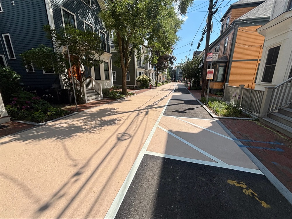 A sunny view of Fairmont Avenue in Cambridge with traditional houses on both sides and a distinctive light beige pavement in the center, featuring pedestrian lines and a bicycle lane symbol.