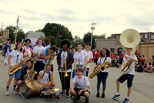 A group of high school students stand in a parking lot with their large brass wind instruments