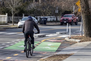 A photo from the Phase 1 project area shows a person on a bike traveling in the two-way bike lane, separated from the vehicle lane with concrete curbs. On the opposite side of the street, two cars are parked.