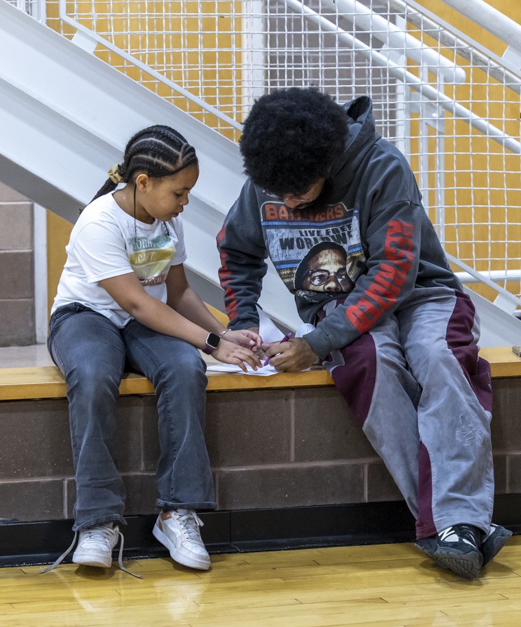 A Cambridge Youth Programs (CYP) staff member guides a youth through a reading activity. Both are seated on a bench in an indoor basketball court.
