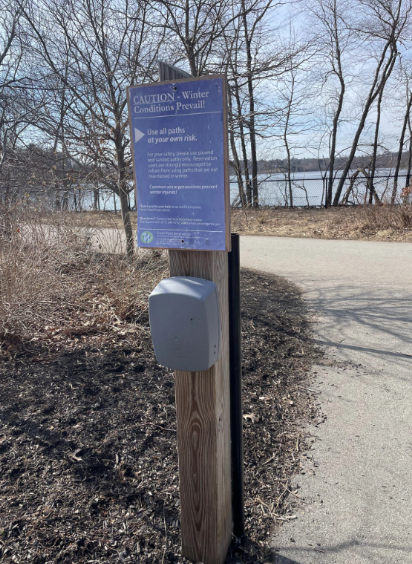 A grey bicycle counter, that looks like a box, mounted on a wooden post at Fresh Pond.