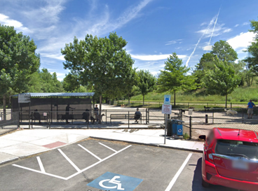 A sandy dog park with a few agility structures at Danehy park. The photo is taken from the angle of the parking lot, with a red car in the lower left hand corner.