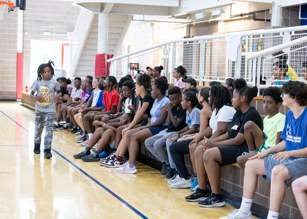 Young Cambridge community members sitting along the bench at the Frisoli Youth Center gym, with one youth attendee speaking to the seated attendees