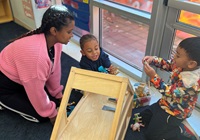 A participant in the Early Childhood Education Career Training Program kneels to play with two preschoolers during her internship at a Cambridge preschool as part of the program.