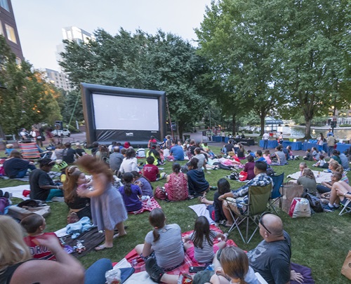 People gathered on the lawn of a park to watch an outdoor movie on a theater-sized screen