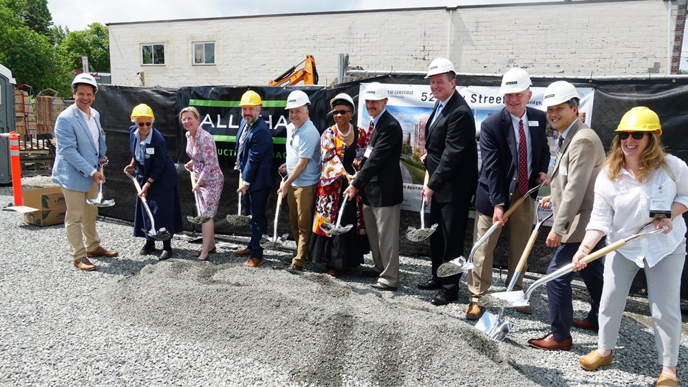 A group of eleven people wearing hard hats and holding shovels participate in a groundbreaking ceremony at a construction site. They are standing on a gravel surface with a backdrop featuring construction fencing and a partially visible sign.