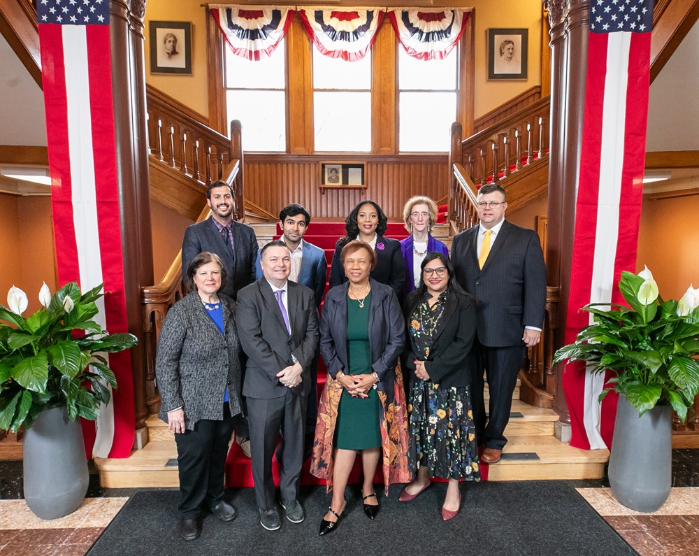 The eight members of the Cambridge City Council posing for a photo in front of the main staircase in City Hall. There's a pillar on each side of the staircase, each flanked with a long, rectangular iteration of the American flag and a green plant in front. Half-circle American flags hang from the windows in the background.