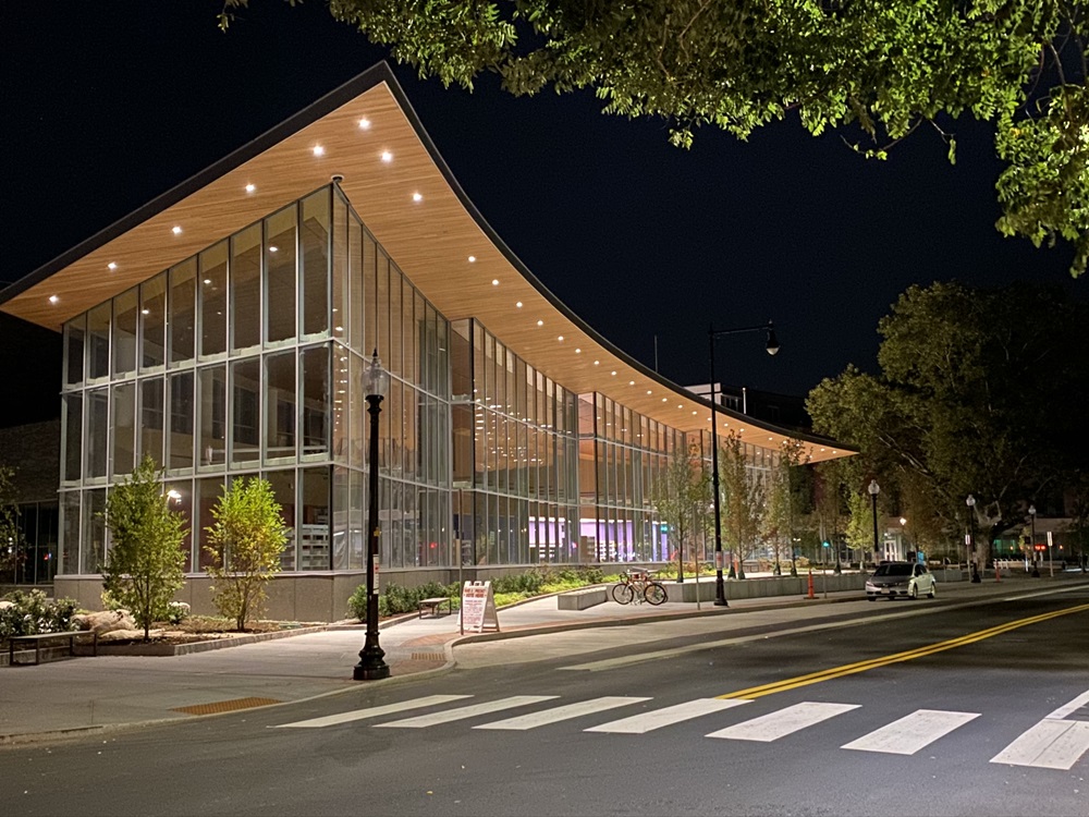 The Valente Library Branch, a modern building with large glass walls and a curved, sloping roof illuminated at night, is located on a tree-lined urban street. Pedestrian crossing lines are visible in the foreground.