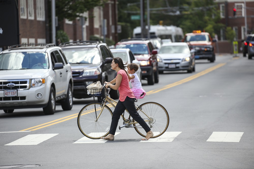 A mother crosses a Cambridge street walking her bike and holding her child on her back