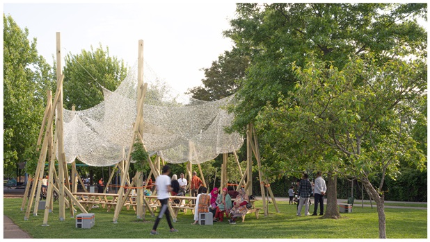 a temporary shade structure made of wood and fabric with a long communal table filled with people standing and seating under it