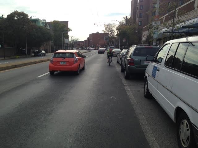 Picture of a person on a bycicle in a very narrow area between parked cars and traffic