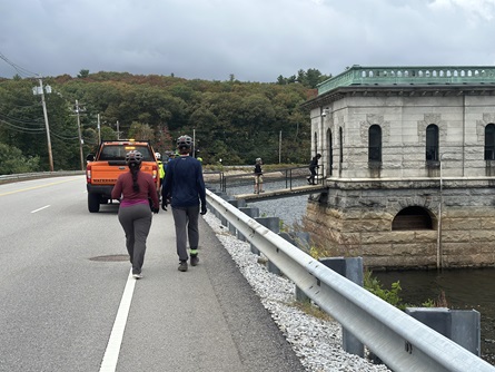 Two riders walk along the winter street dam. An orange watershed truck and the winter street gatehouse are in the background.