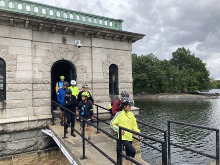 People walk along a bridge connecting a road to the winter street gatehouse at Hobbs Brook Reservoir. The gatehouse is white granite.