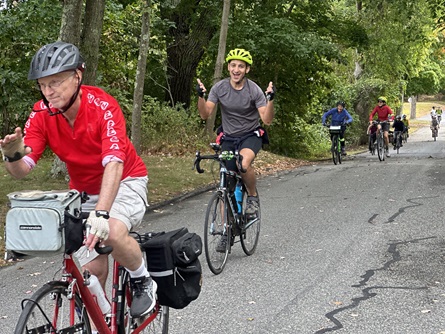 People smile and wave at the camera while biking along a road.