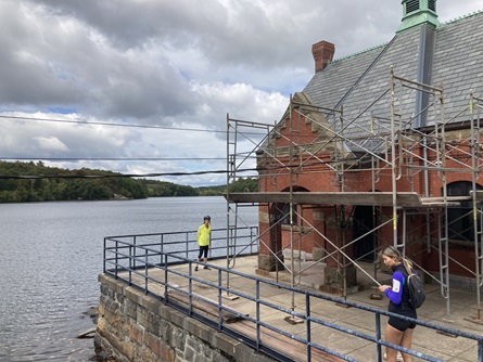 Two people stand on the concrete pad outside of the Stony Brook Reservoir gatehouse. The gatehouse is red brick, surrounded by construction scaffolding.