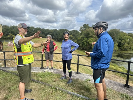 Riders stand in the grass along Stony Brook Reservoir dam and ask staff members questions.