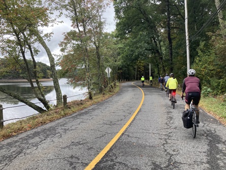 Riders seen from the back as they bike along a two-lane road next to a reservoir.