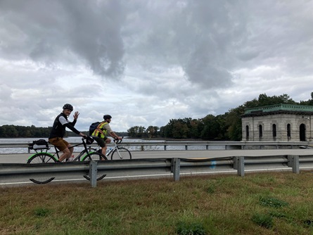 Two riders bike along a dam. A white gatehouse and the reservoir are in the background.