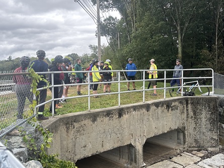 People gather behind a fence as a staff member talks to them. Below the people is a culvert and a rocky spillway for a dam.