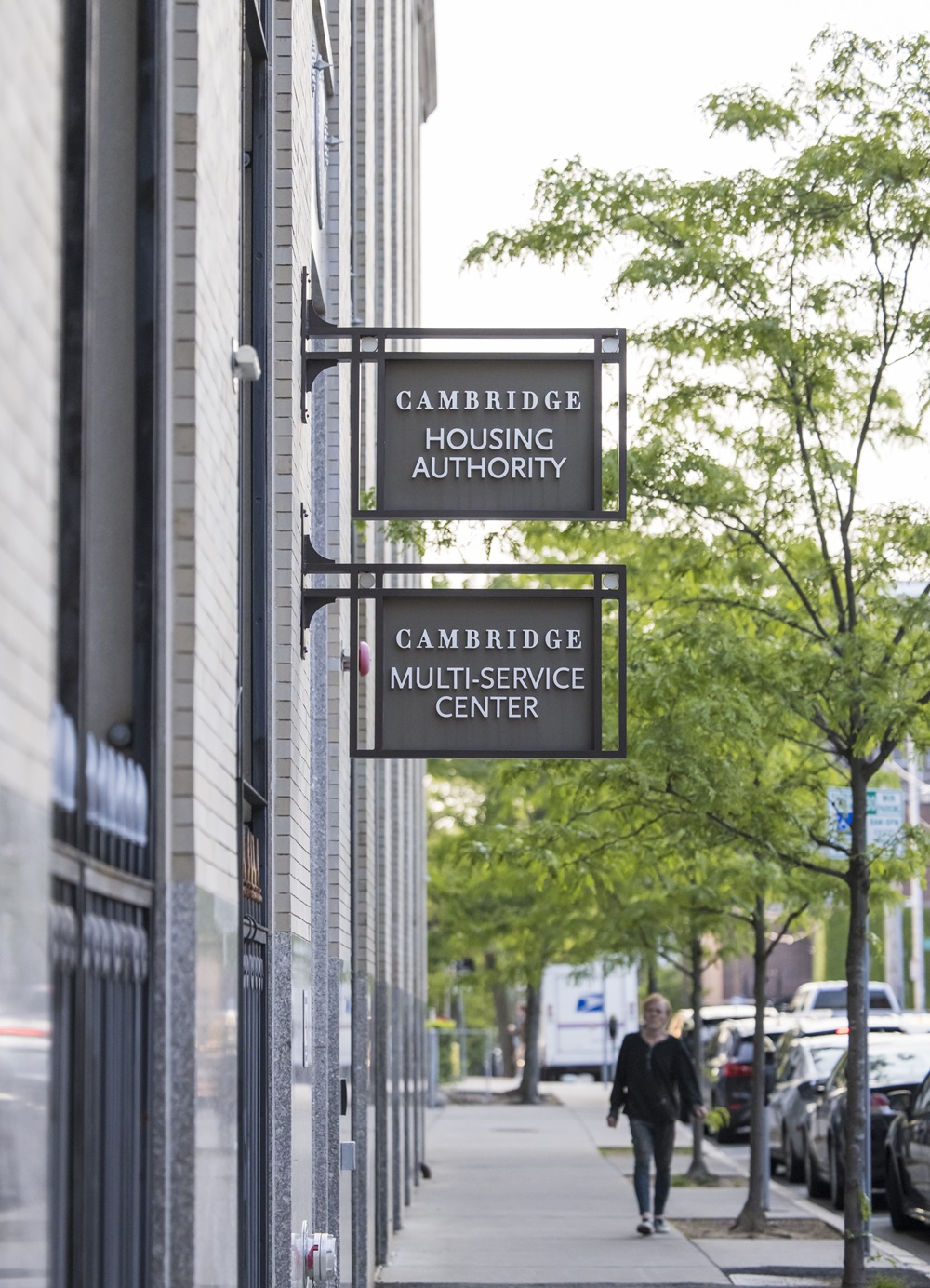 Signs for the Cambridge Housing Authority and Cambridge Multi-Service Center hang on a city building, with a person walking on the sidewalk beside parked cars.
