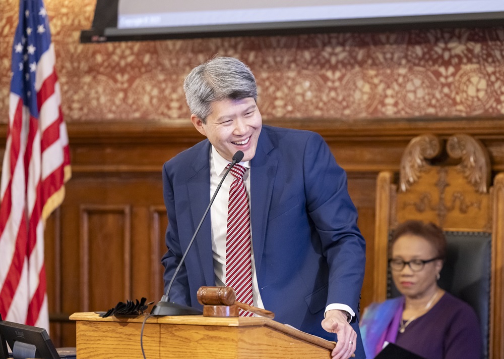 Cambridge City Manager Yi-An Huang is smiling while speaking at a podium in the Sullivan Chamber at Cambridge City Hall.